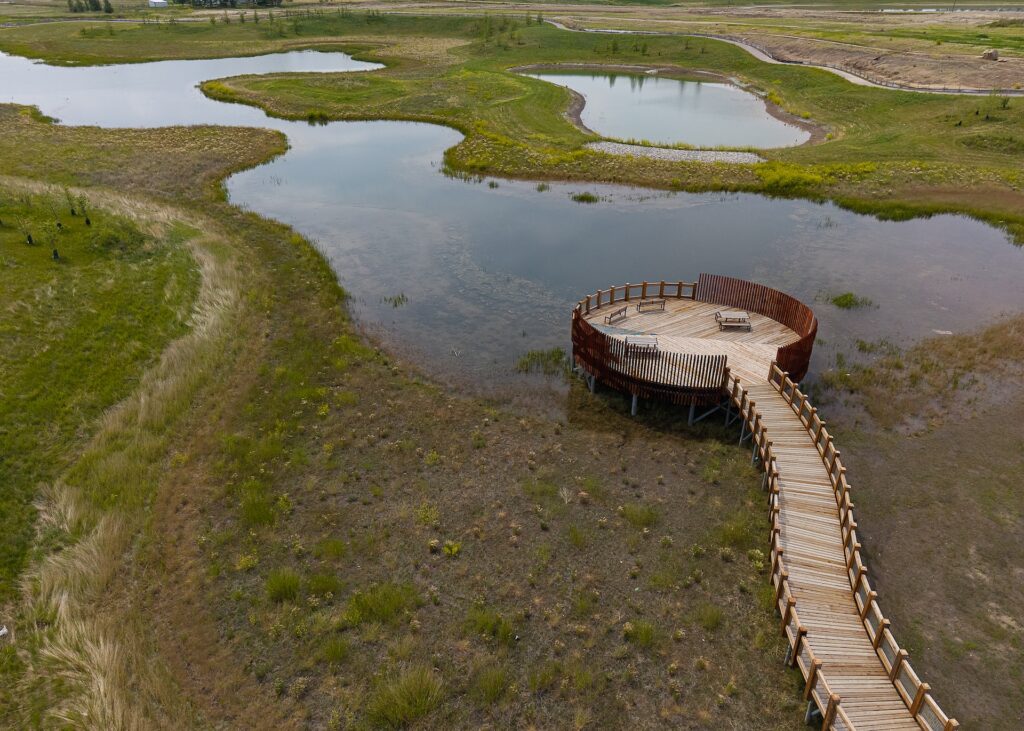 Aerial view of a circular wooden viewing platform with benches, connected to shore by a winding boardwalk, surrounded by marshland and shallow water.