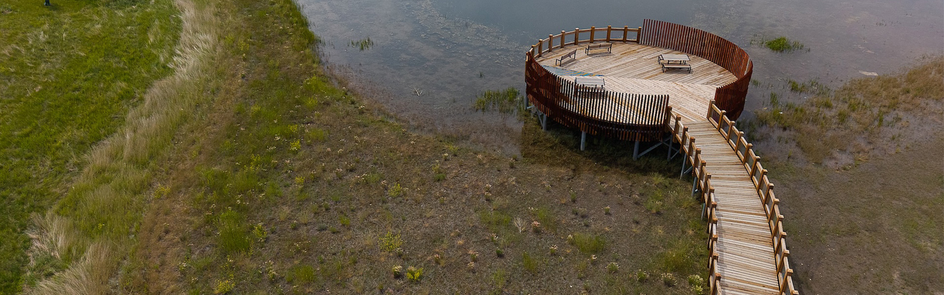 Aerial view of a circular wooden viewing platform with benches, connected to shore by a winding boardwalk, surrounded by marshland and shallow water.