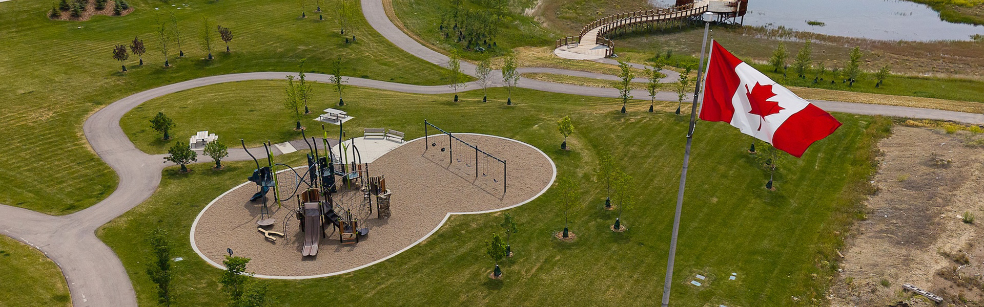 Aerial view of a playground with slides, climbing structures, and swings, set in a grassy park with walking paths, a large Canadian flag, and the wetland boardwalk in the background.